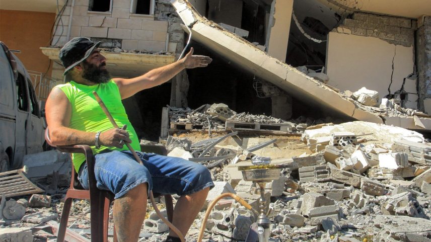 Ali Mallah fuma narguilé nos escombros de seu prédio em Ain Qana, no sul do Líbano, destruído por ataque aéreo israelense (Foto: Mahmoud Zayyat/AFP)