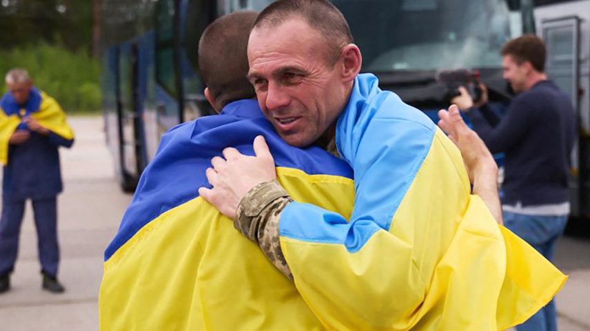 Prisioneiros de guerra ucranianos (POWs) envoltos em bandeiras nacionais se abraçam após uma troca de abraços em um local não revelado, em meio à invasão russa na Ucrânia (Foto: UKRAINIAN PRESIDENTIAL PRESS SERVICE / AFP)