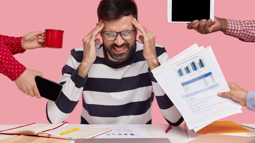 Homem de marketing sobrecarregado esfrega as têmporas, precisa se concentrar e trabalhar no relatório financeiro, focado para baixo, segura uma caneca de café quente, papéis com gráficos, touchpad e celular, posa em ambiente interno. (Foto: Freepik)