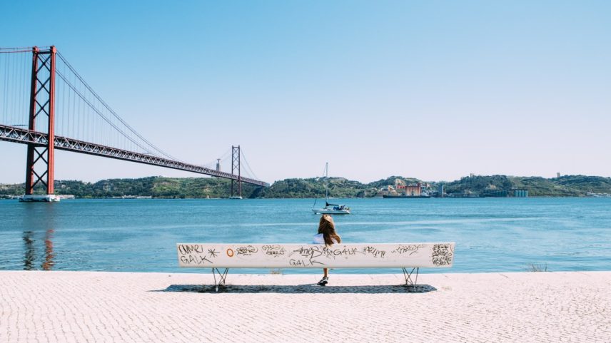 Mulher sentada em um banco à beira do Rio Tejo, em Lisboa (Foto: Jason Briscoe/Unsplash/Divulgação)