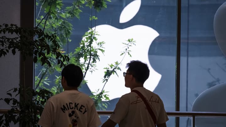 Dois jovens homens estão dentro de um shopping em frente a um grande logo iluminado da Apple visto através de uma janela em Chongqing, China, em 4 de junho de 2025. (Foto: Cheng Xin | Getty Images)