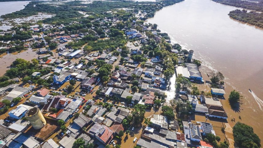 Vista aérea de área residencial às margens de rio atingida por inundações, com as águas ocupando o leito das ruas por entre as casas, que se concentram na parte inferior da imagem, enquanto na superior aparecem áreas verdes tomadas pelas águas (Foto: USP Imagens)