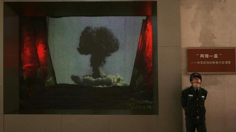 A screen fixed to a wall shows a video of a mushroom cloud. A soldier in a formal dress uniform stands beside the screen with his hands clasped behind his back as he leans against the wall.