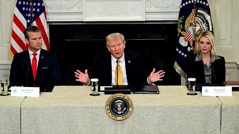 Hegseth, Trump, and Bondi sit at a table with the presidential seal affixed to it. All our dressed in business formal attire. Trump has his arms outstretched as he speaks.