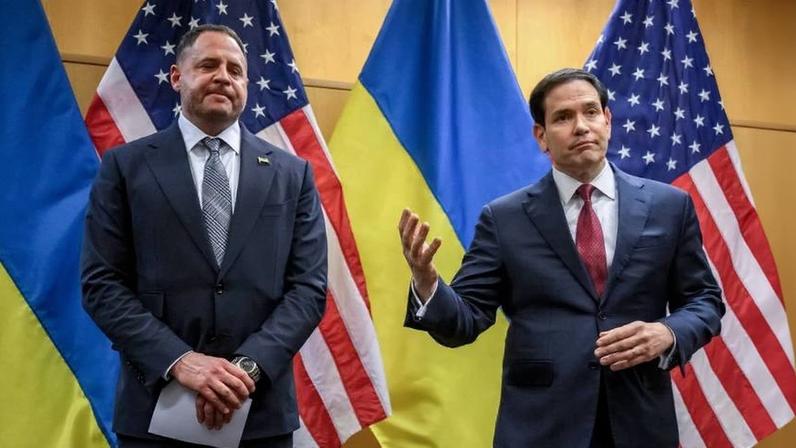 Yermak and Rubio stand side by side in front of U.S. and Ukrainian flags. Yermak is looking downward, while Rubio gestures with his right arm in what appears to be a slight shrugging motion.