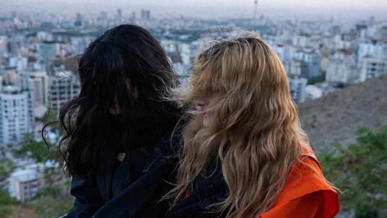 Two women with long hair are seen in profile with their hair covering their faces. Behind them are the buildings of a city.