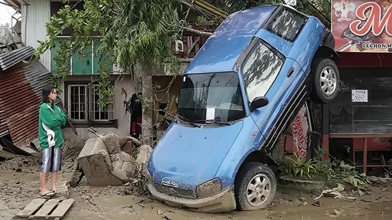 Getty Images A young woman in a green and white jacket looks on forlornly as a blue car hangs from a tree, with its rear end in the air, having been lifted by floods during Typhoon Rai in the Philippines in 2021