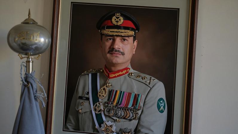 A framed portrait photograph covered with broken glass tilts to the right on an off-white wall, showing Munir wearing a formal army uniform. A silver flag pole with a pike on top stands in the foreground.