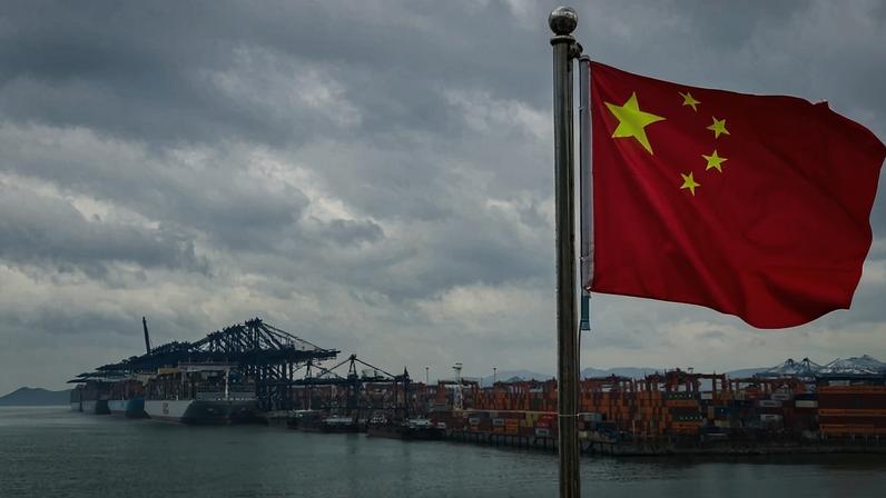 Chinese national flag is seen in the foreground with container ships, cranes, and stacked shipping containers.