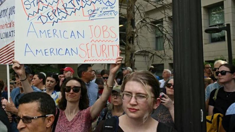 In a crowd of people standing outside, one woman wearing sunglasses holds aloft a hand-written sign that reads in part, "State Department: Thank you for your service."