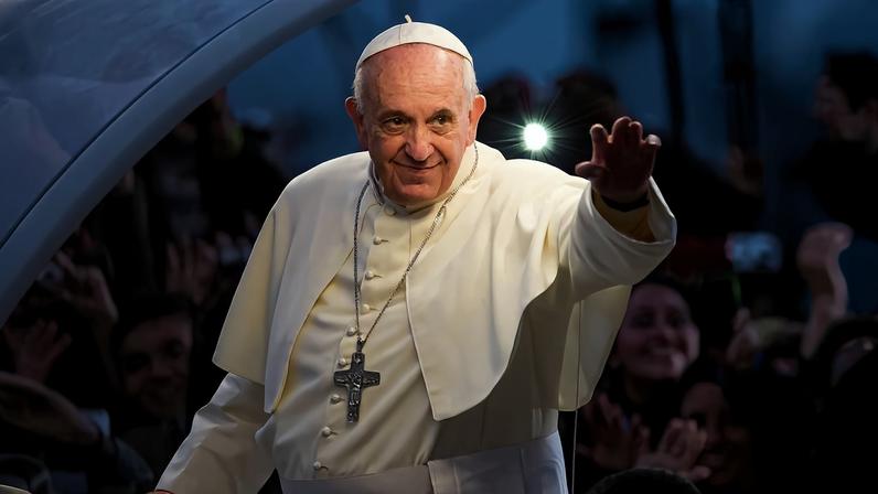 Pope Francis, dressed in white, waves at a crowd of people at dusk. A lash of light shines above his shoulder.