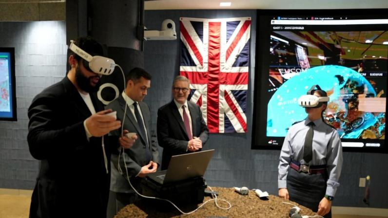 Starmer and other personnel stand around a circular table, trying out white AI-powered goggles and handheld instruments. The British flag is on bright display on the wall behind them.