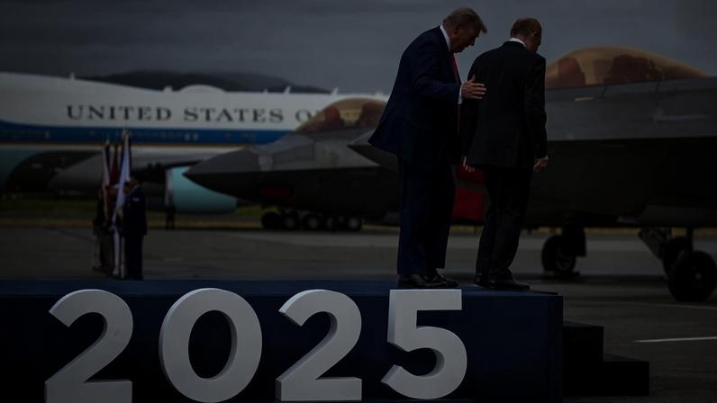 Trump and Putin stand on a blue stage with the letters 2025 affixed to the front of it in large white figures. Trump's hand is on Putin's back to guide him as he walks onto the steps off the stage. In the background, jets and Air Force One are parked on an airport tarmac.
