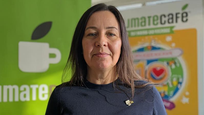 Jess Pepper, a dark-haired woman in a black sweater and a Climate Café pin badge, stands in front of a the café signage which depicts a coffee mug and a colourful 10-year anniversary graphic.