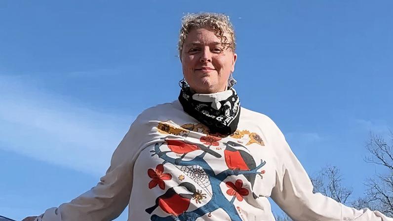 Paradox Mercer looking at camera. She has blonde curly hair. She is wearing a white long-sleeve top with an image of a blue branch with red and dark blue birds on it. There is blue sky in the background with trees. She has a bandanna round her neck.