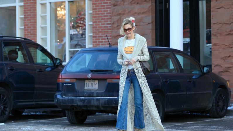 Bella Hadid walking outdoors in Aspen, wearing a sheer lace duster coat, a graphic T-shirt with a smiley face, flared jeans, and pointed-toe boots.
