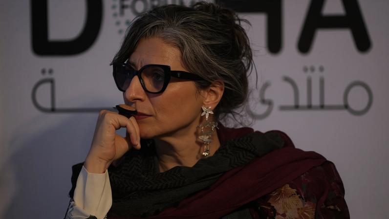 Albanese rests her hand against her face thoughtfully as she listens to someone out of frame speak. The Doha Forum logo can be seen on a large screen in the background.