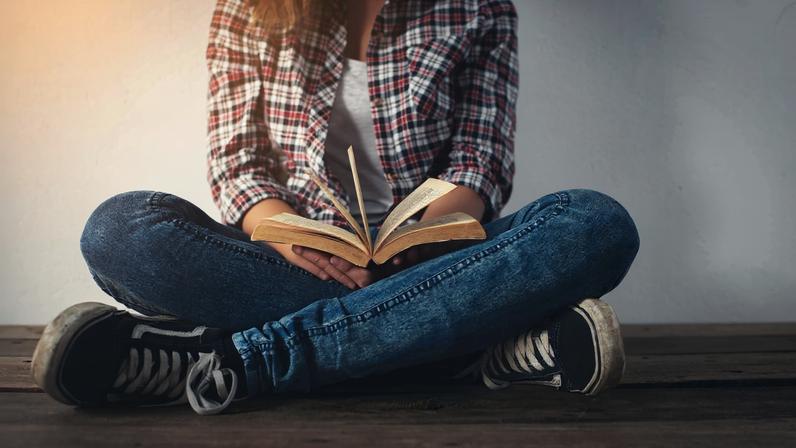 Young hipster woman holding open book sitting on wooden floor.