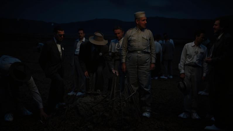 In this black-and-white photo, a group of men lean over a twisted metal object protruding from the ground in a desert landscape. Most of the men wear military uniforms or suits and ties.