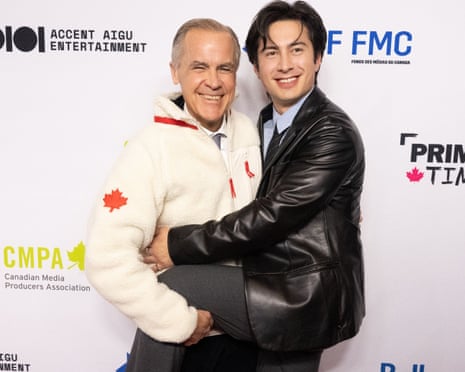 Hudson Williams, in a leather jacket and smart clothes, puts his leg around Mark Carney, who is wearing a white fleece and holding onto Hudson's leg