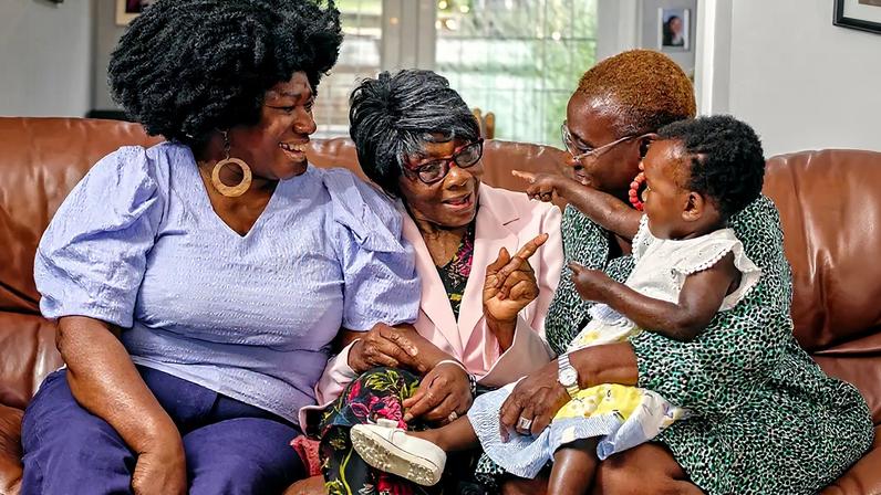 Fotografia de mulheres negras de idades diferentes sentadas no sofá com mãe idosa e bebê.