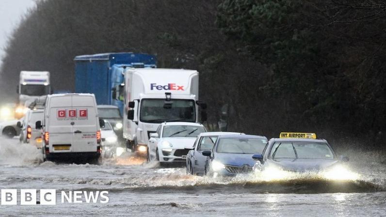 Cars drive through heavy floods on the roads in Northern Ireland. There are several cars and lorries driving through deep water. They hae their headlights on as the light is low