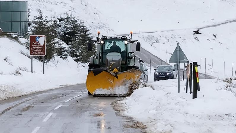 PA Media A tractor with a yellow snow plough attached to the front drives along a road, with a black SUV behind it. The edges of the road and the surrounding hillsides are covered in snow.