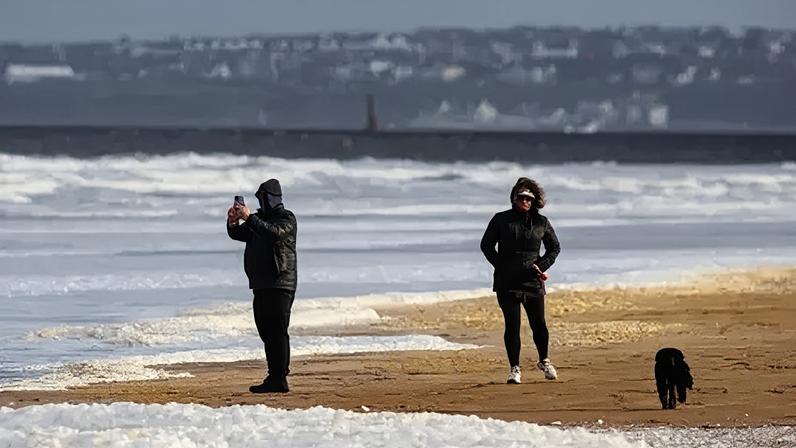 PA Media People wrapped in jackets and hats walk along a beach.