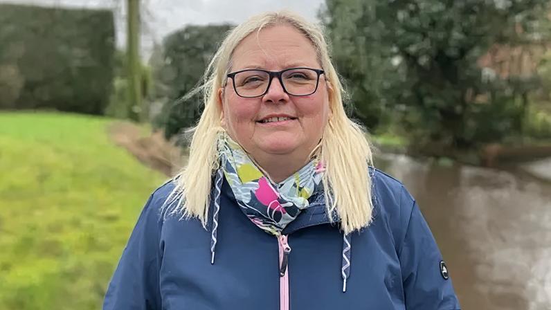 Debbie Sinclair, a woman with blonde hair, is wearing a blue coat and a scarf with a green and pink pattern. She is standing outdoors with a flooded road behind her.