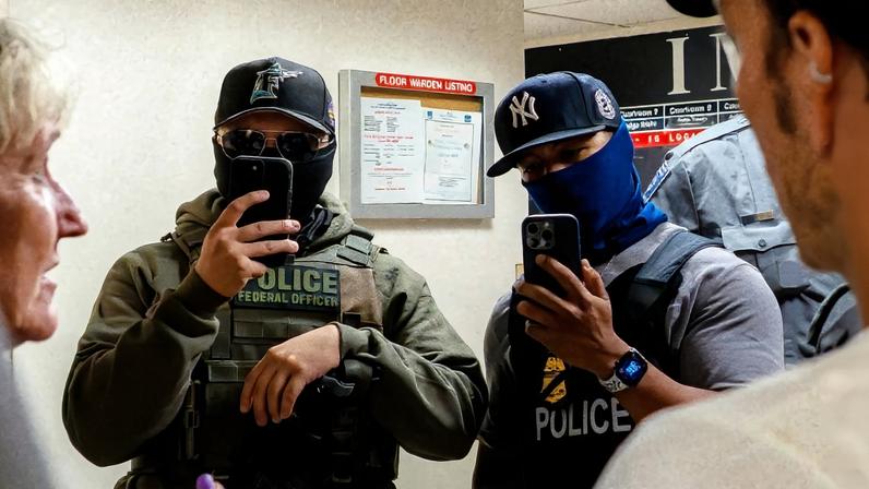 Two ICE agents film the press using smartphones in the hallway outside the immigration court at 26 Federal Plaza in New York City.