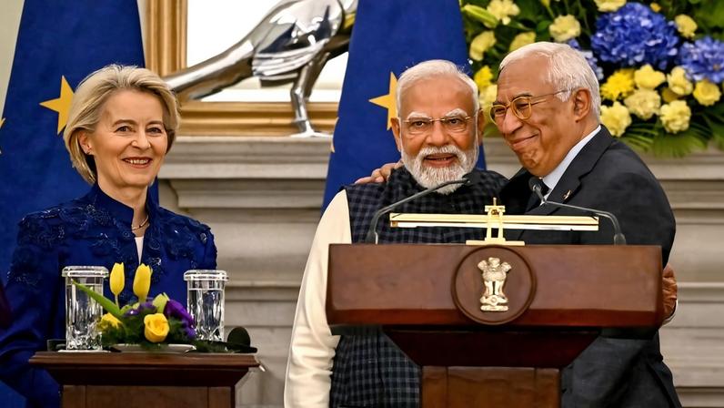 Von der Leyen stands at a podium beside Modi and Costa, who are embracing each other, as they pose for the camera.