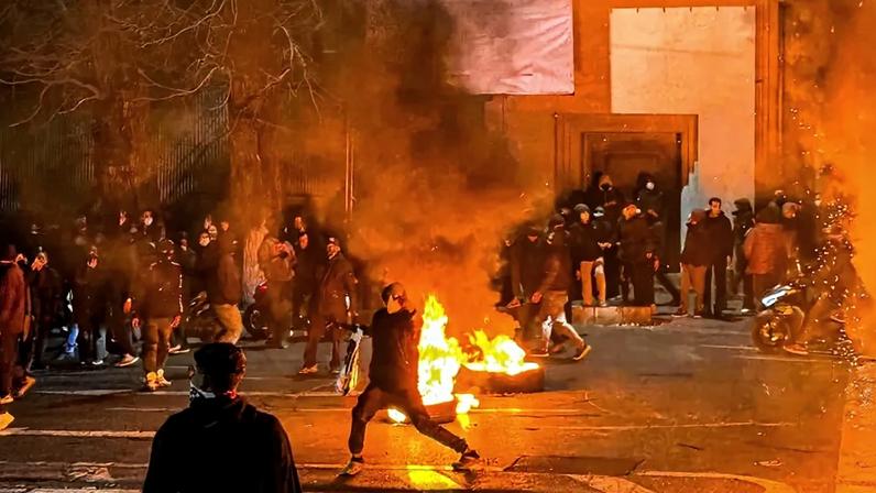 Iranians block a street in Tehran amid burning objects and heavy smoke.