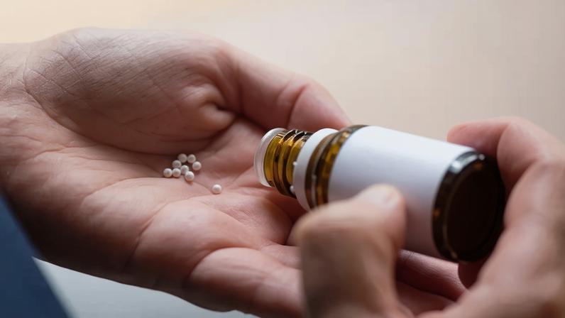 Uma pessoa derramando pílulas homeopáticas (glóbulos) de um frasco de vidro na palma da mão.