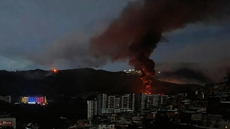 A plume of smoke and fire rises above tall buildings and in front of hills in Caracas against a night sky.