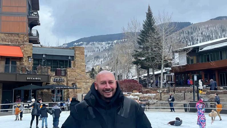 Jasen Kaplan smiling on an outdoor ice rink with buildings and snow-covered mountains in the background.