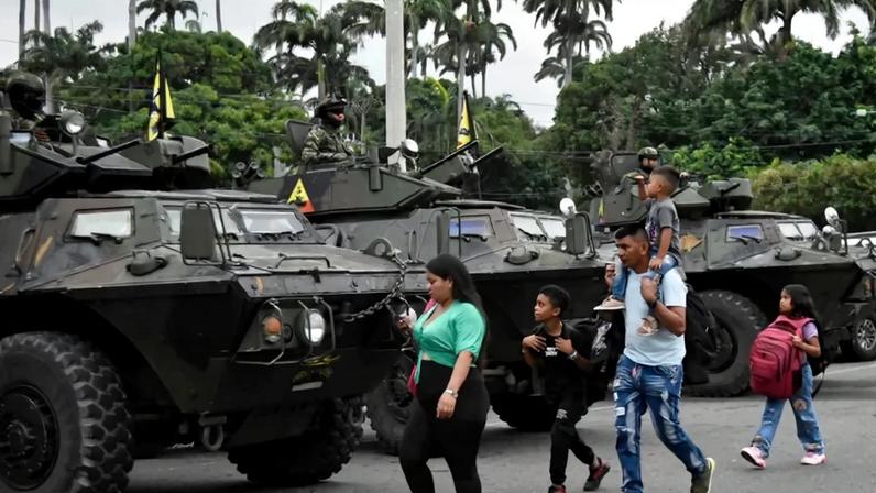 A woman and man walk with three children, one on the man's shoulders, past a row of armored military vehicles with soldiers in combat gear. The children carry backpacks. Palm trees are seen in the distance.