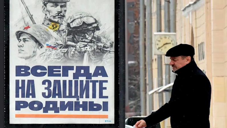 A man in black clothes and a black hat walks past a billboard depicting three eras of soldiers with lettering in Cyrillic.