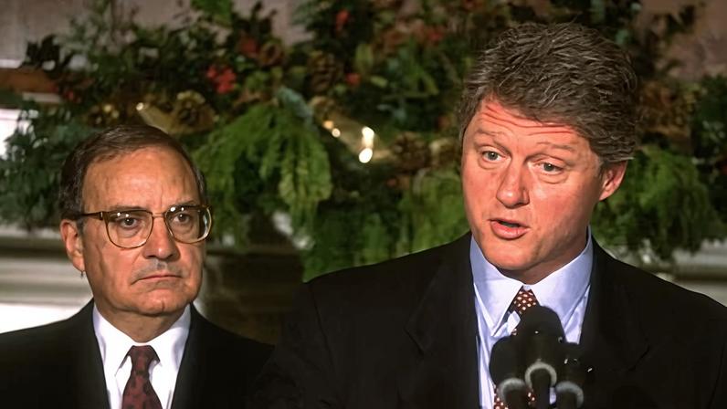 Envoy George Mitchell stands behind Bill Clinton as the former president speaks into a microphone