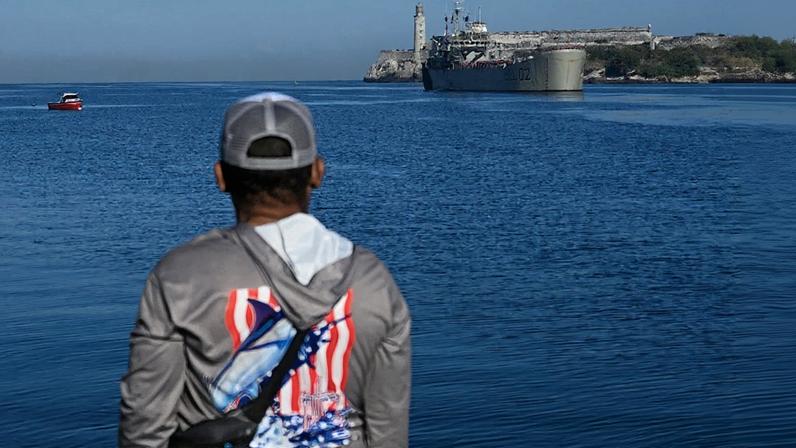 A man in a gray baseball cap and gray sweatshirt with an American flag on the back looks out at a Mexican navy ship approaching from across Havana Bay.
