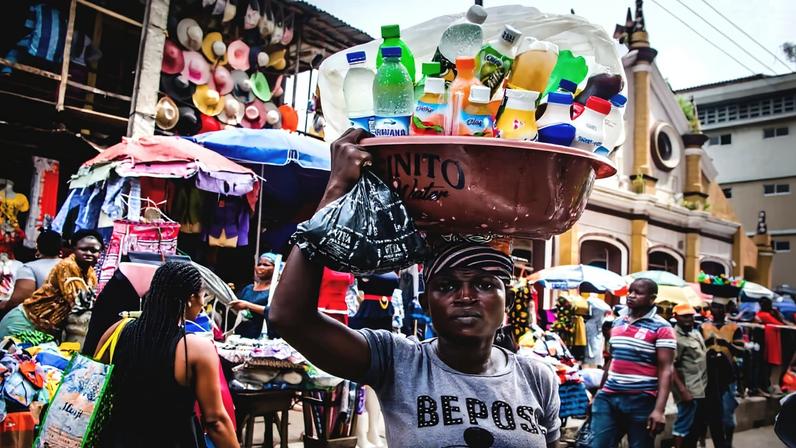 A woman standing in a street carries a basin filled with a colourful variety of plastic bottles while in the background there are busy stalls and shoppers in the street. Image by Elliot Ovadje via Flickr.