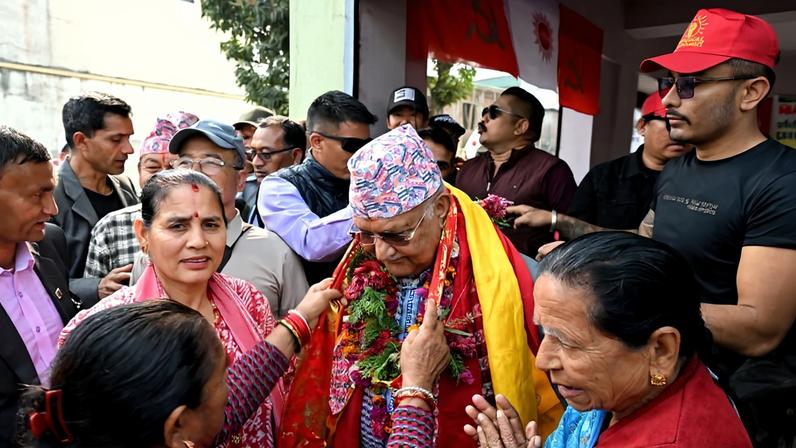 Oli, an older Nepalese politician, stands among a crowd of supporters as a few of them place a garland over his head.