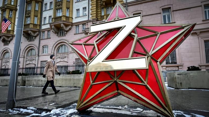 A large sculpture of a red-and-gold star with a gold Z affixed to the front stands in front of a building. A person bundled in a winter coat and hat walks by in the background.