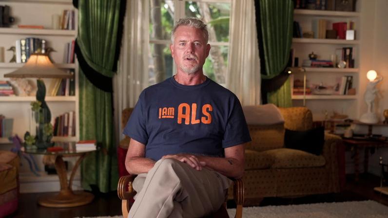 Eric Dane sitting in a living room, wearing a blue t-shirt with "I AM ALS" written in orange.