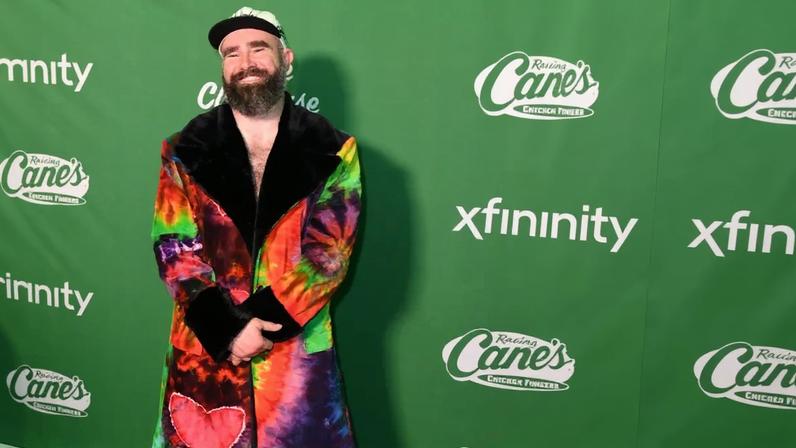 Jason Kelce smiling in a colorful tie-dye coat with a red heart patch, light-wash jeans, and white sneakers, standing on a green astroturf carpet with white yard lines.