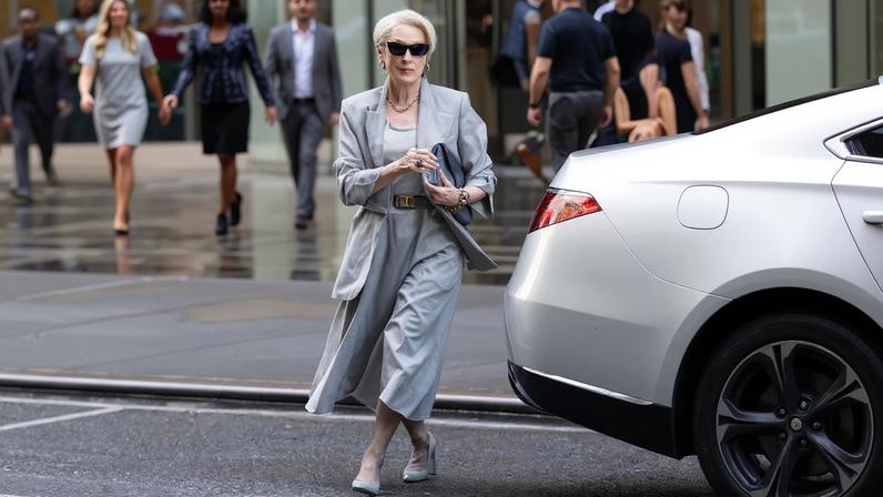 Meryl Streep in character as Miranda Priestly, wearing a light gray skirt suit and sunglasses, stepping out of a silver car.