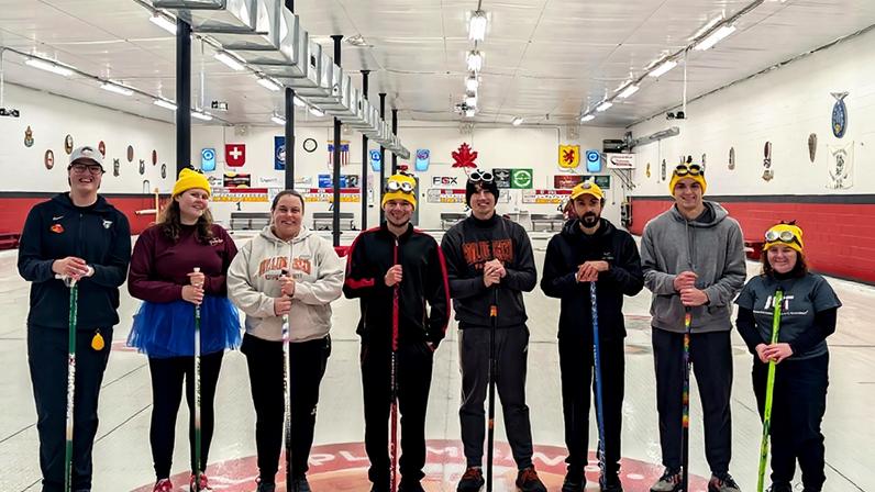 8 members of MIT's Curling team pose standing in a hockey rink.
