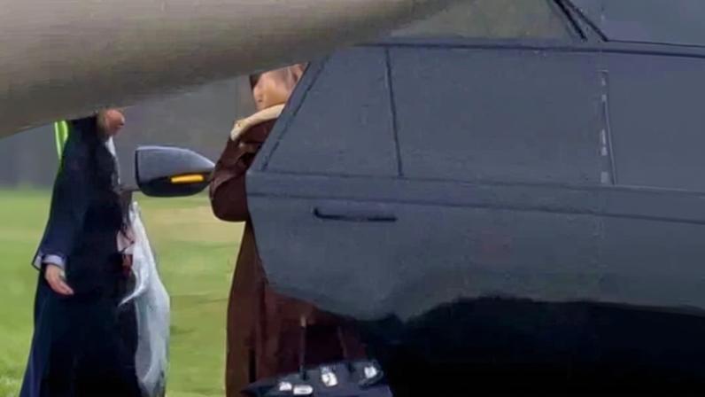 Kim Kardashian, wearing a brown coat and black sunglasses, holding a black Hermes Birkin bag, stands next to a car and a private jet.