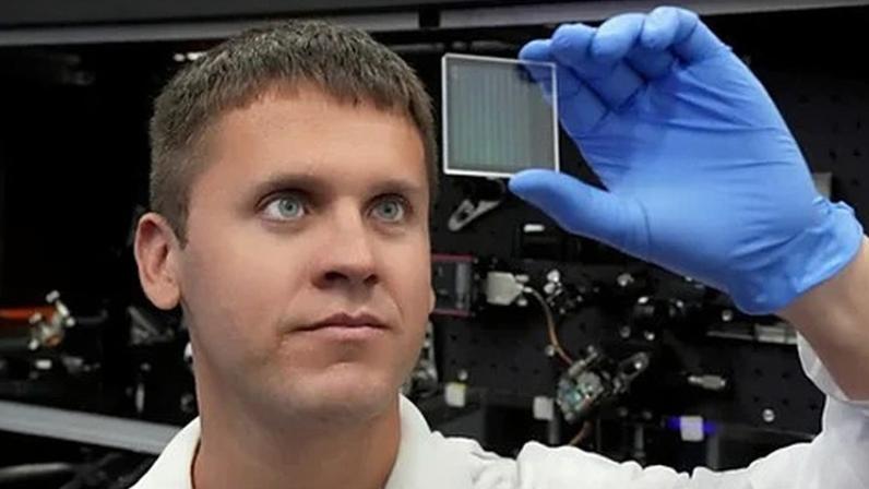 SPhotonix Ilya Kazansky, CEO of Sphotonix wearing a white lab coat and blue latex gloves holds up a small square glass plate with grid markings on it to see through it (Credit: SPhotonix)