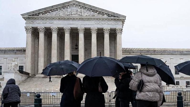 People stand under umbrellas while facing the steps in front of the U.S. Supreme Court building under an overcast sky.