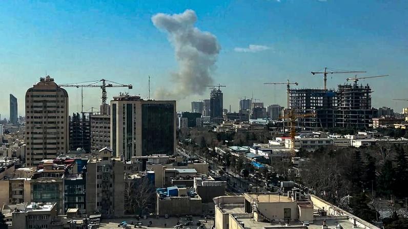 A plume of smoke rises in a blue sky behind a cityscape.